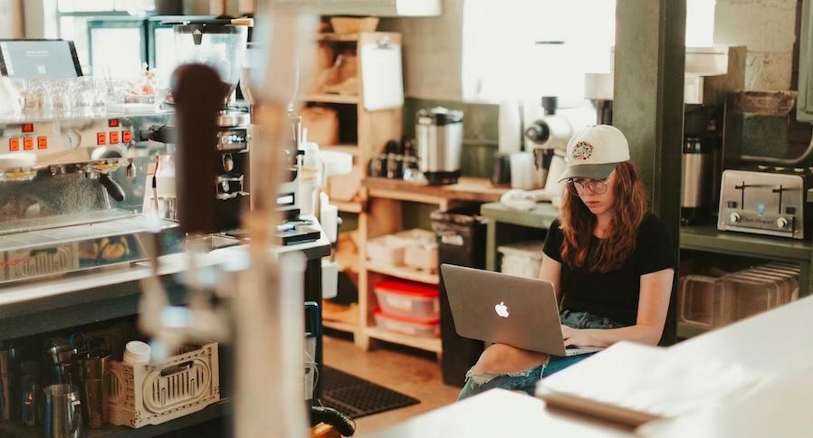 A hip, middle-aged, female business owner working on a laptop behind the counter of her cafe